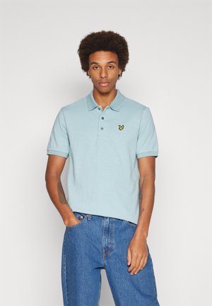 Polo shirtazzurro in misto cotone con colletto, tre bottoni e un logo ricamato giallo sul lato sinistro del petto. Indossato con jeans in denim blu.
