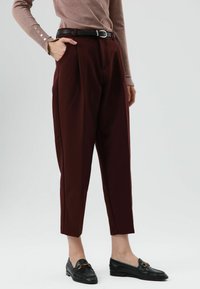 Bordeauxrode, op maat gemaakte broek met plooien, hoge taille, taps toelopende pijpen en een zwarte riem. Gedragen met zwarte loafers, gestructureerde stof, gladde afwerking.