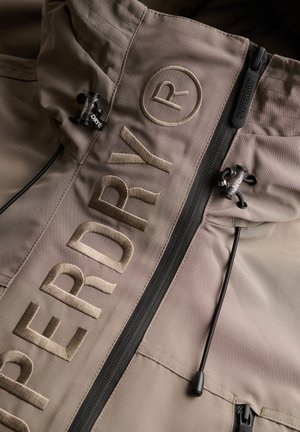 Giacca beige con grande scritta ricamata "SUPERDRY®", cerniera nera con tiretto "SUPERDRY" e coulisse nere regolabili sul cappuccio.
