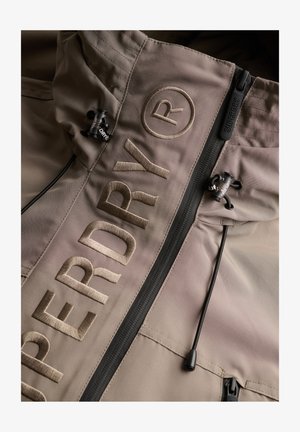 Veste beige avec grand texte brodé "SUPERDRY®", fermeture éclair noire avec tirette "SUPERDRY" et cordons noirs réglables à la capuche.