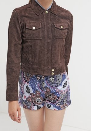 Personne portant une veste en daim marron avec des boutons pression et un short à motif cachemire multicolore sur un fond uni.