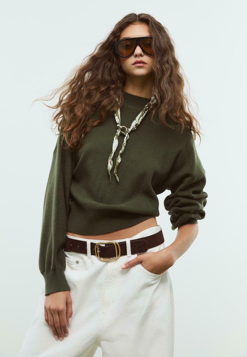Una donna con lunghi capelli ricci indossa occhiali da sole oversize marroni, un maglione verde oliva, pantaloni bianchi, una cintura marrone e una sciarpa a fantasia, posando con fiducia.