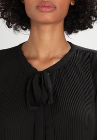 Blouse noire plissée avec un col rond et un détail de lien au col. Le tissu a une texture lisse avec de subtiles lignes verticales.