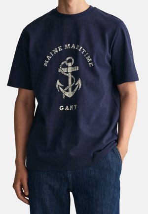 Homme portant un t-shirt bleu marine avec un motif d’ancre et de corde blanc et le texte « Maine Maritime GANT », associé à un jean bleu.
