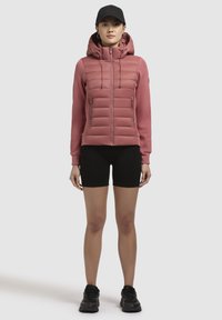 Gewatteerde roze jas met een hoge kraag, rits aan de voorkant en dubbele zijzakken. Gecombineerd met zwarte shorts en schoeisel, tegen een neutrale achtergrond.