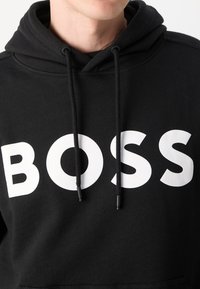 Jeune personne portant un sweat à capuche noir avec le texte BOSS blanc sur la poitrine et des cordons noirs pendants.