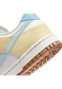 Zapato con un diseño de múltiples materiales que incluye paneles blancos, amarillos y azul claro, superficies texturizadas y una suela de goma marrón con un patrón de tracción.