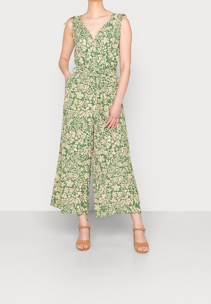 Jumpsuit verde con motivo floreale, scollatura a V, gambe larghe e dettaglio annodato in vita. Il tessuto sembra leggero e dalla consistenza texture.
