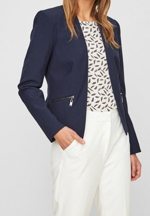 Femme portant un blazer bleu marine avec des poches zippées, un haut blanc à motifs et un pantalon blanc taille haute, une main dans la poche.