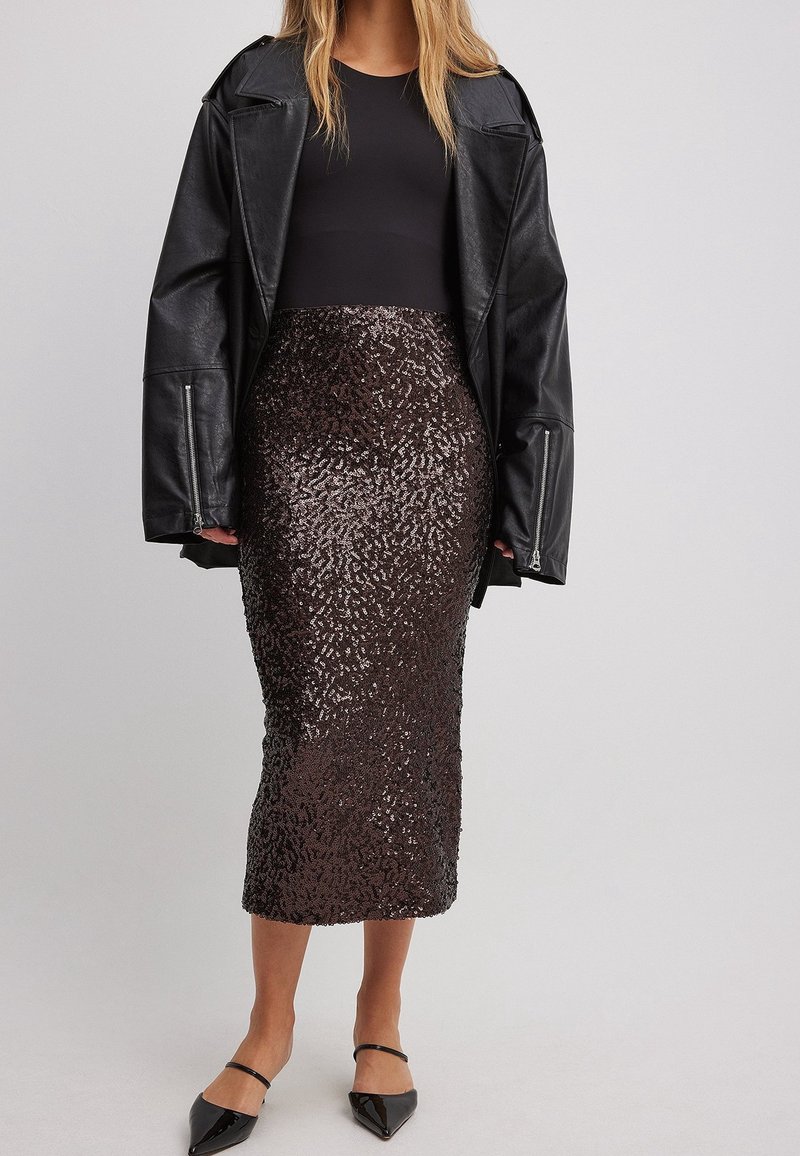 Femme portant une veste en cuir noire sur un top noir, associée à une jupe à sequins marron foncé longueur genou et des talons pointus noirs.