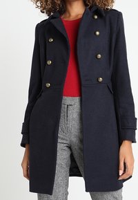 Cappotto doppiopetto in misto lana blu navy con bottoni dorati, colletto a punta e tasche laterali, abbinato a una maglia rossa e pantaloni a quadri.