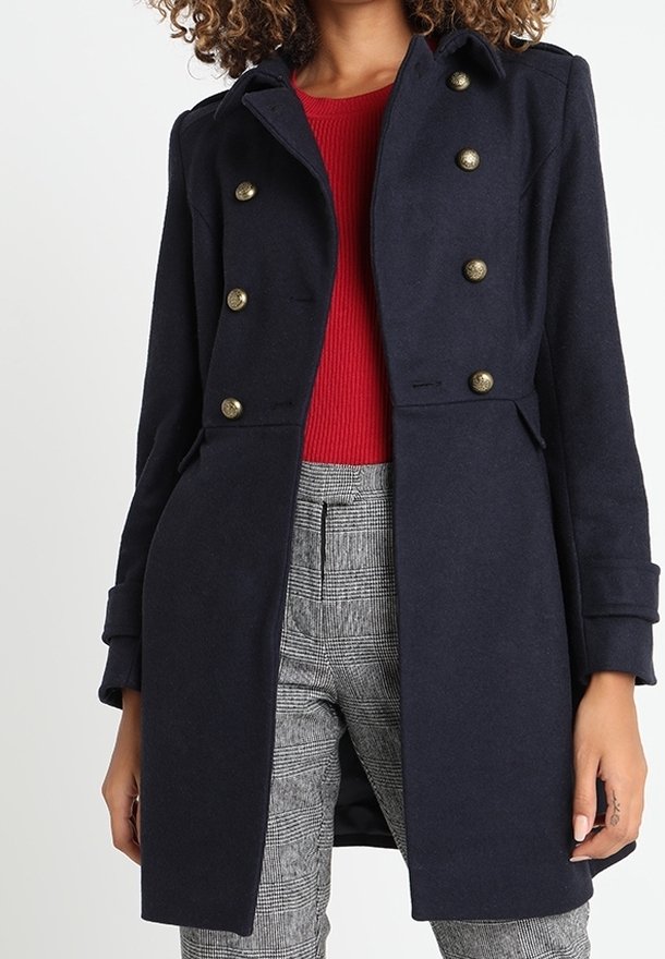 Cappotto doppiopetto in misto lana blu navy con bottoni dorati, colletto a punta e tasche laterali, abbinato a una maglia rossa e pantaloni a quadri.