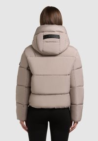 Beżowa kurtka puchowa z kapturem, wyróżniająca się pikowanym wzorem i wyraźną naszywką z logo na plecach. Gładki, błyszczący materiał o luźnym kroju.
