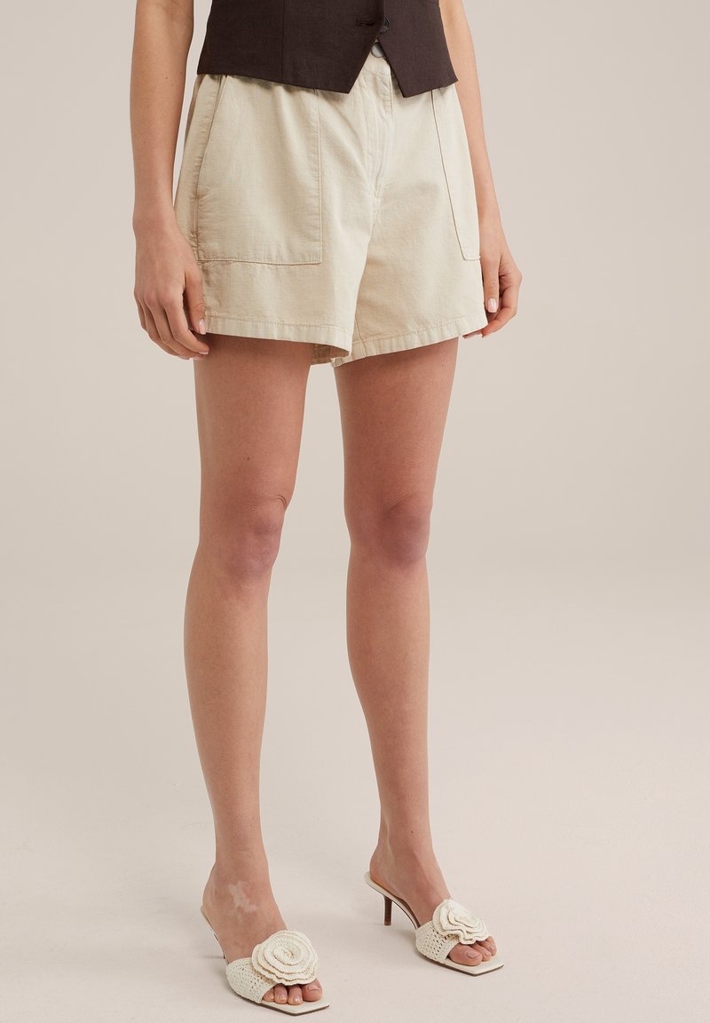 Shorts en coton beige clair avec deux poches avant, associés à un gilet marron ajusté et des sandales à talons blanches avec un accent en forme de rose.