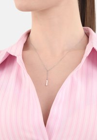 Silver kedja med tre rosa pärlor hängande vertikalt, bärs med en ljusrosa randig skjorta.