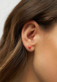 Pendiente en forma de corazón rojo, asegurado con herrajes dorados, llevado en el lóbulo, con textura suave y acabado pulido.
