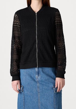 Kvinde iført en sort blonderjakke med lynlås og gennemsigtige mønstrede ærmer samt en blå denims nederdel med forlommer.