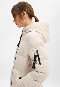 Beżowa kurtka puchowa z kapturem, czarnymi zamkami błyskawicznymi i bocznymi kieszeniami. Teksturowany materiał z wypukłymi sekcjami zapewniającymi ciepło.