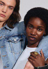 La veste en jean présente un lavage bleu clair, un col classique et des éléments en métal. Le modèle porte une chemise blanche et une bague texturée dorée sur une peau foncée.