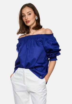 Frau trägt eine blaue schulterfreie Bluse mit langen Ärmeln und weiße Hose, Hände in den Taschen, vor einem schlichten hellen Hintergrund.