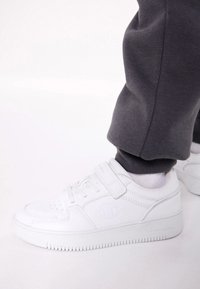 Baskets blanches avec une tige synthétique lisse, une sangle velcro, une semelle texturée et un logo sur le côté. Assorties à un pantalon de survêtement gris foncé.