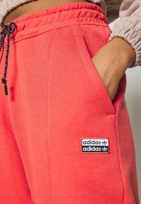Coral athletic joggers with elastic waistband, drawstring, side pockets, and black Adidas logo patch. Soft, smooth texture.