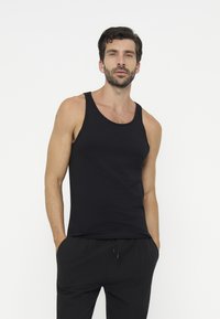 Uomo con capelli scuri e barba che indossa una canotta nera senza maniche e pantaloni neri, con le mani nelle tasche, su uno sfondo chiaro e uniforme.