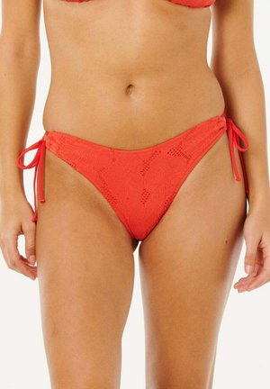 Kvinde iført rød blomsterbroderet bikiniunderdel med snorebind ved siderne, vist fra talje til øvre lår mod hvid baggrund.