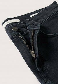 Jean en denim noir plié avec une fermeture éclair argentée partiellement ouverte et un bouton noir à la ceinture montrant les détails des poches et des passants de ceinture.