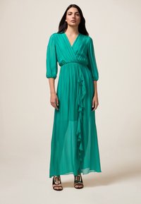 Rochie lungă din chiffon verde smarald cu un design înfășurat, cu volane în față și mâneci trei sferturi. Dispune de o talie strânsă și o siluetă vaporoasă.