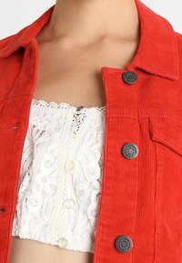 Giacca di corduroy rossa con colletto e bottoni metallici, indossata sopra una maglietta bianca in pizzo con chiusure a bottoni e motivi intricati.