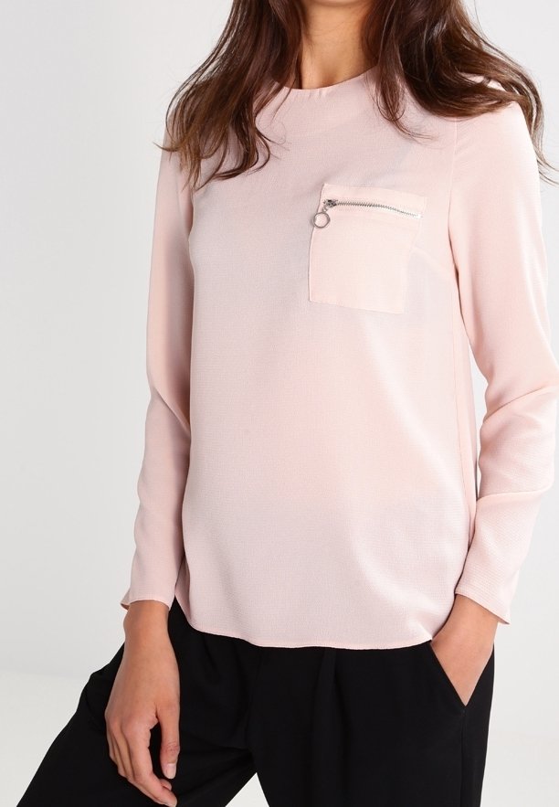 Haut à manches longues rose clair avec une finition texturée et une poche plaquée unique ornée d'un détail en zip métallique, porté avec un pantalon foncé.