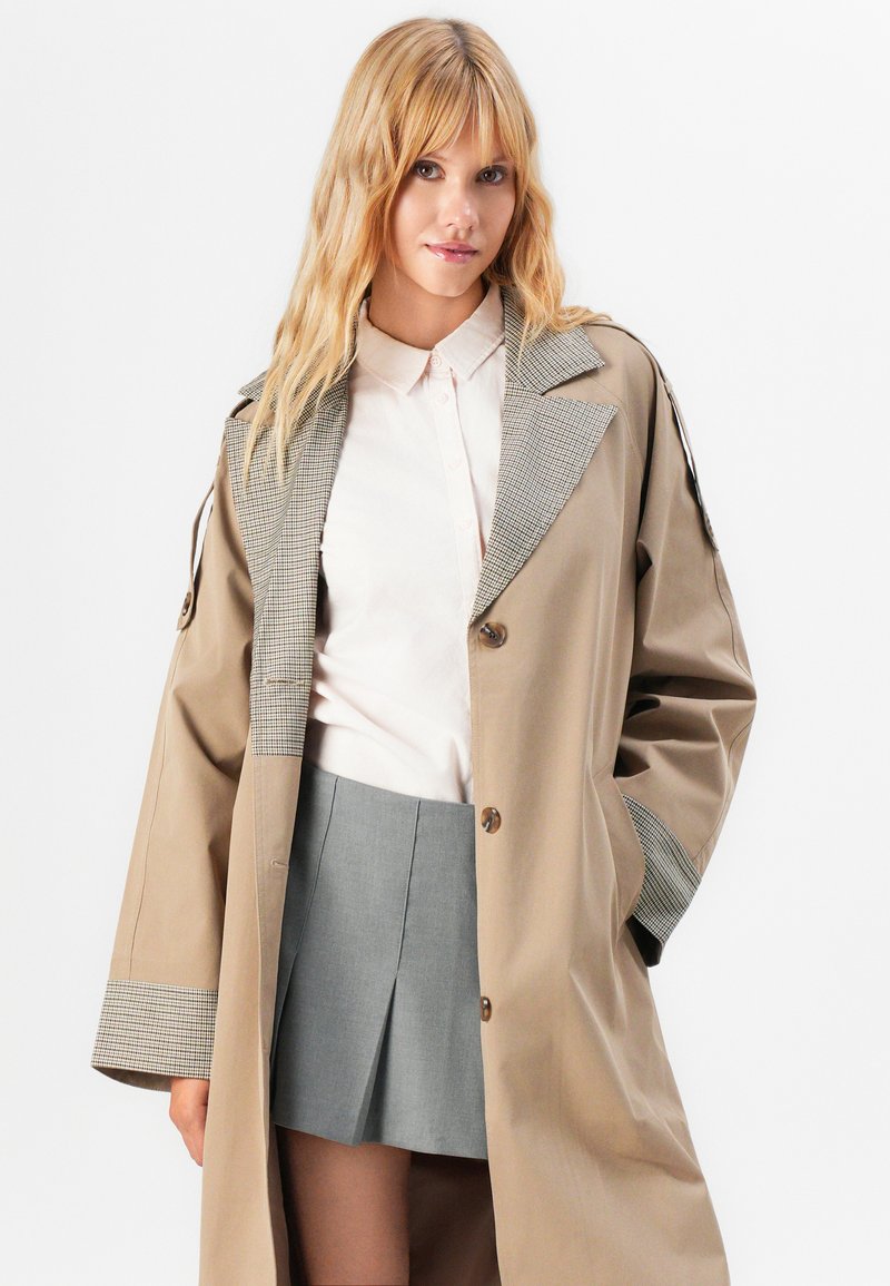Femme blonde portant un trench-coat beige avec col et poignets à carreaux, une chemise blanche à boutons, et une jupe plissée grise, se tenant devant un fond blanc.