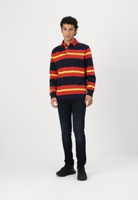 Langærmet polo t-shirt i marineblå med vandrette røde og gule striber. Har krave, tre-knaps lukning og logo-detalje. Mørke jeans og sorte sneakers.