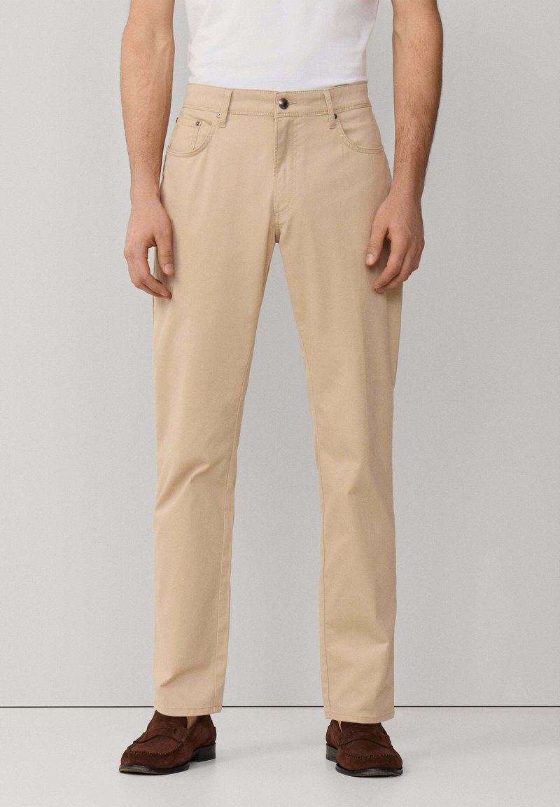 Mann trägt beige, gerade geschnittene Hose, weißes Hemd eingesteckt und braune Wildleder-Loafer, steht vor einem schlichten hellen Hintergrund.