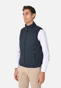 Gilet senza maniche di colore blu navy realizzato in materiale leggero e liscio. Presenta un colletto alto, una zip frontale e due tasche laterali con accenti arancioni.