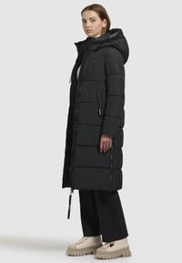 Lange zwarte gewatteerde jas met een capuchon, doorstikt ontwerp, rits aan de voorkant, zijzakken en een tweekleurige textuurafwerking. Gepaard met stevige beige laarzen.