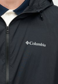 Μαύρο μπουφάν Columbia με φερμουάρ μπροστά φορεμένο πάνω από λευκό πόλο πουκάμισο με σκούρο μπλε γιακά.