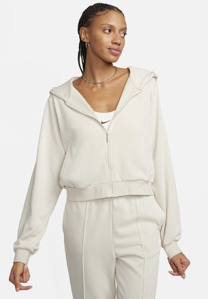 Cremefarbener Hoodie aus weichem Stoff, mit Reißverschluss, übergroßer Kapuze und gerippten Bündchen. Wird mit passenden cremefarbenen Jogginghosen getragen.