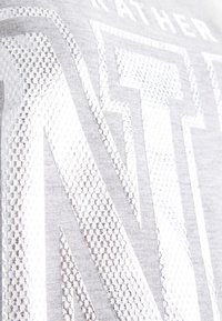 Stříbrné grafické tričko s texturovaným designem a hexagonálními vzory na světlém šedém pozadí. Doplňuje ho odvážné typografické prvky.