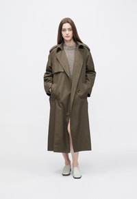Olivgrön trenchcoat med dubbelknäppt design, midjebälte, sidofickor och medellång skärning, kombinerad med vita slip-on-skor.