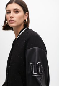 Čierna bomber bunda so softshellovými rukávmi, ozdobená nýtovanými detailmi a koženými rukávmi, s korálkovým logom "LC" na ľavom ramene.