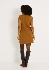 Vestido de pana sin mangas en marrón con forma ajustada y cremallera en la parte posterior. Combinado con botas de cuero marrón hasta la rodilla. Textura suave.