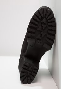 Black shoe with a textured rubber sole, featuring deep treads and an angular design. The upper has a smooth finish and is sleek in shape.