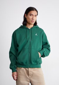 Grøn zip-up hoodie lavet af bomuld. Den har en kængurulomme, justerbar hætte med snore, og et lille hvidt logo på brystet.