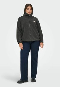 Szara bluza polarowa na zamek z wysokim kołnierzem, zestawiona z ciemnymi dżinsami i czarnymi butami na platformie. Na piersi znajduje się małe logo.