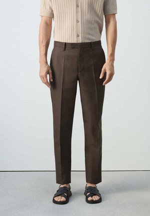 Hombre con pantalones marrones a medida, camisa de punto beige de manga corta y sandalias negras abiertas, de pie contra un fondo sencillo.