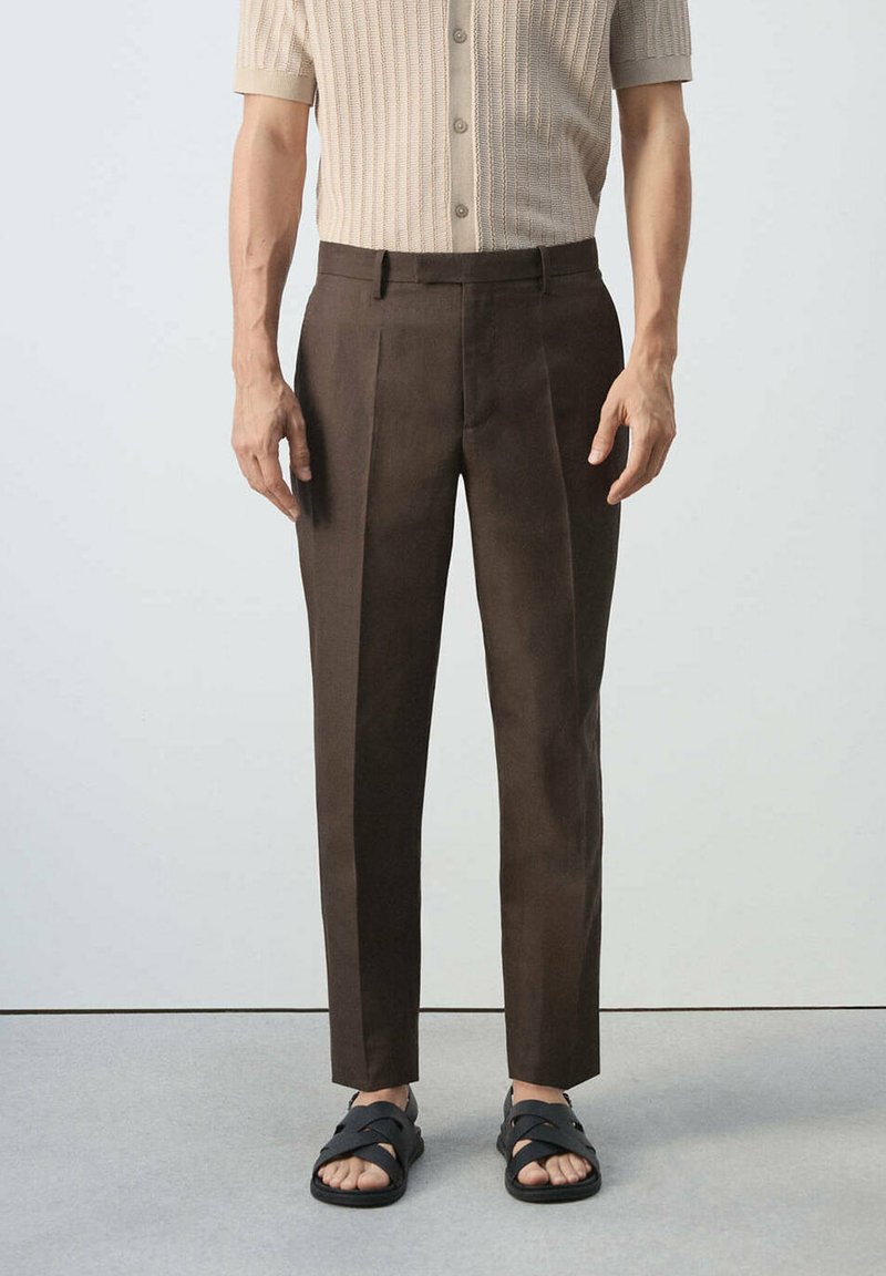 Homme portant un pantalon marron sur mesure, un pull beige à manches courtes et des sandales noires ouvertes, debout devant un fond uni.