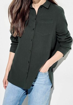 Kvinde iført en mørkegrøn langærmet skjorte med brystlomme, kombineret med lyseblå jeans, hånd i lommen.