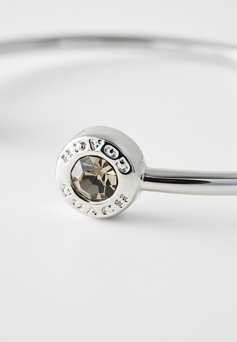 Pulsera de plata con un colgante redondo que presenta una piedra facetada y el texto "COACH" en relieve. Acabado metálico suave y diseño limpio y minimalista.